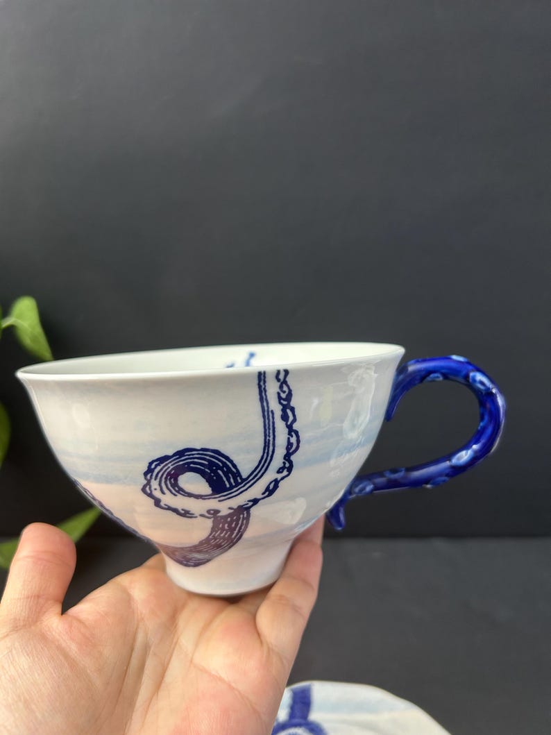May include: White ceramic teacup with a blue octopus tentacle design. The cup has a blue handle and a blue and white painted sky design. The cup is held in a hand against a dark background.