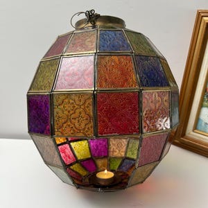 May include: A spherical lantern constructed from multi-colored, textured glass panels held together by a metal frame. The glass features a repeating embossed pattern in shades of red, blue, green, yellow, and purple. A small lit tea light candle sits inside.