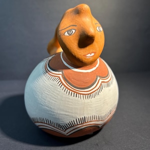 May include: A handmade ceramic jug with a human face. The face is painted in shades of brown, with white-rimmed eyes and a small mouth. The body is white with brown and black geometric patterns. The jug has a rounded shape and a handle.