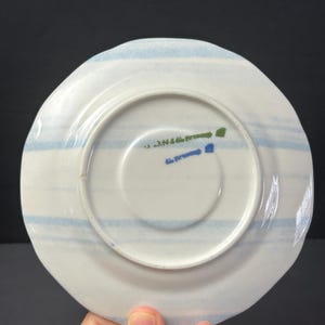 May include: White ceramic saucer with light blue horizontal stripes. The underside features a central white circle and text in green and blue. The saucer has a slightly irregular edge.