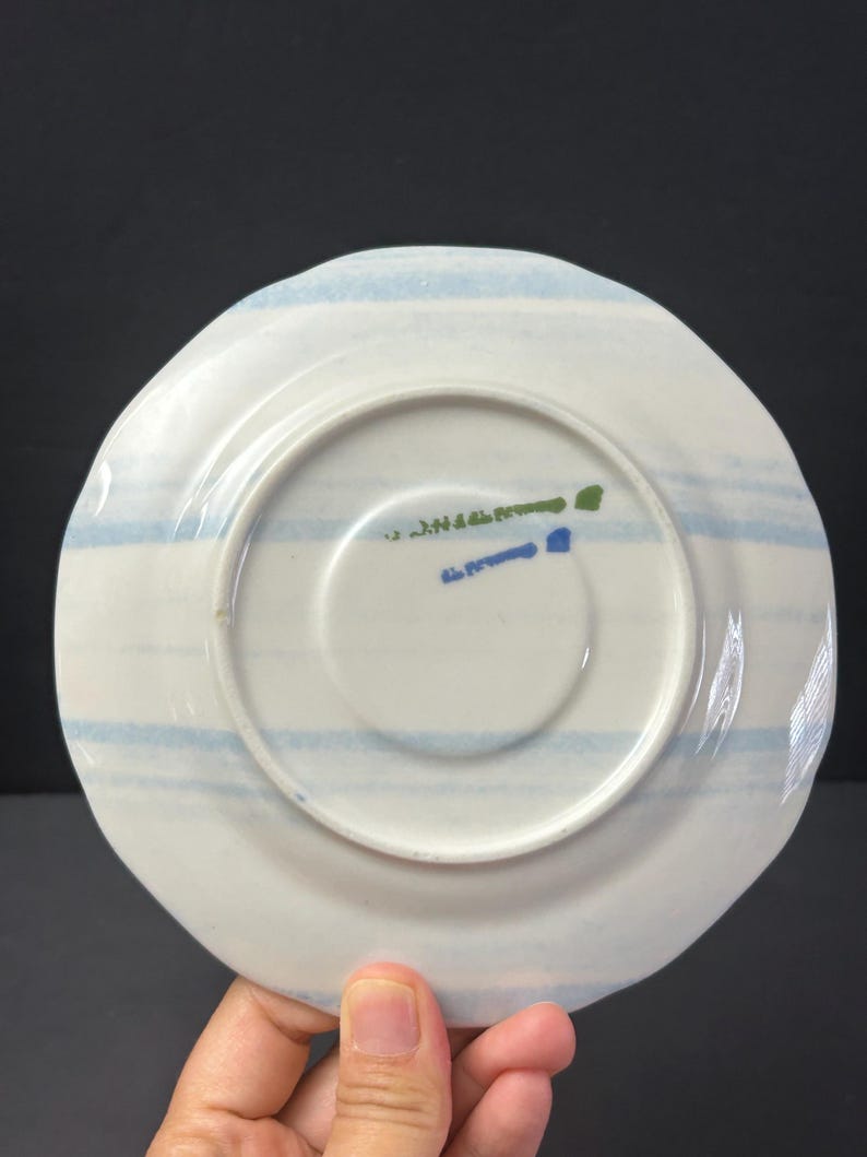May include: White ceramic saucer with light blue horizontal stripes. The underside features a central white circle and text in green and blue. The saucer has a slightly irregular edge.