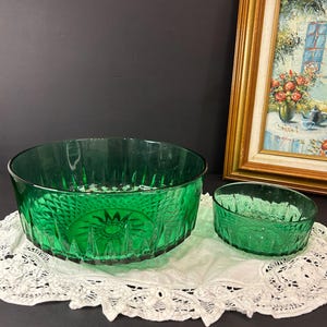May include: Two emerald green glass bowls with a textured pattern sit on a white lace doily. The larger bowl is oval, and the smaller bowl is round. A framed painting with a gold frame is in the background.