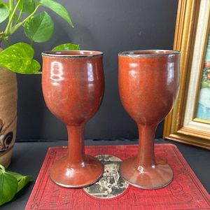 May include: Two handmade, reddish-brown ceramic goblets with a speckled finish and dark rims. The goblets are displayed on a vintage red book. The stemware has a classic, elegant design, suitable for serving beverages. The background includes a plant and a framed painting.