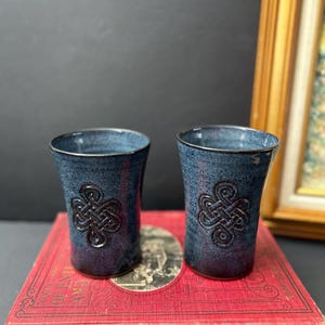 May include: Two cylindrical ceramic tumblers with a speckled blue and purple glaze. Each tumbler features a raised, embossed Celtic knot design on the front. The vessels have a smooth, slightly tapered shape with a dark rim and base.