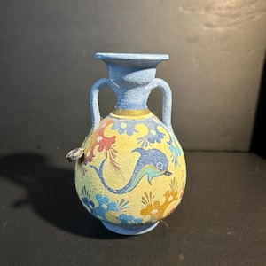 Puede incluir: Un pequeño jarrón decorativo con borde, asas y base de color azul claro. El jarrón tiene un cuerpo amarillo con un diseño de delfín y floral pintado en azul, rojo y dorado. El jarrón mide unos 13 cm de altura.