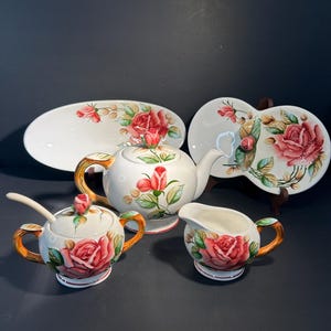 May include: A white ceramic tea set featuring a red rose pattern. The set comprises a teapot, sugar bowl, creamer jug, and two serving plates. The teapot and sugar bowl have orange handles. Plates are oval and heart-shaped.