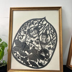 May include: A framed black paper cutout featuring intricate Thai-style figures playing stringed instruments. The artwork is set against a cream-coloured background and enclosed in a rectangular gold-toned frame with detailed filigree patterns.