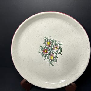 May include: A speckled, off-white ceramic plate with a red rim and a floral design in the center. The flowers are pink, yellow, and red with green leaves. The plate is displayed on a dark brown wooden stand.