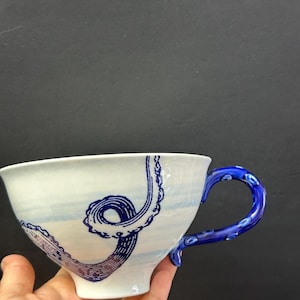 May include: White ceramic teacup with a blue octopus tentacle design. The handle is a vibrant blue with a textured pattern. The cup has a wide rim and a curved shape, perfect for tea or coffee.