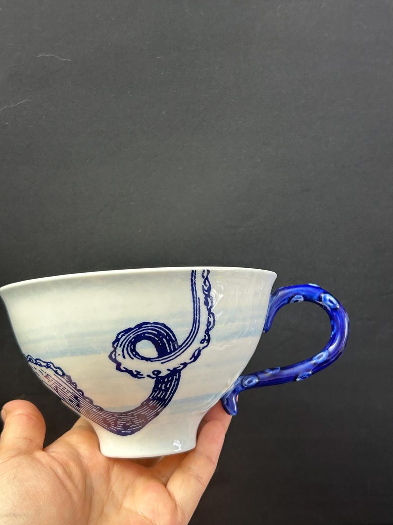 May include: White ceramic teacup with a blue octopus tentacle design. The handle is a vibrant blue with a textured pattern. The cup has a wide rim and a curved shape, perfect for tea or coffee.