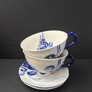 May include: Two stacked white teacups and saucers with blue nautical-themed designs. The cups have blue handles and feature a sailboat illustration. The saucers have matching blue wave patterns.