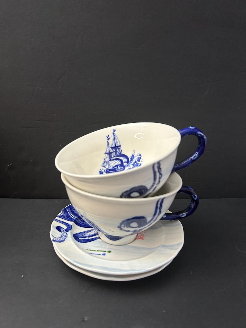 May include: Two stacked white teacups and saucers with blue nautical-themed designs. The cups have blue handles and feature a sailboat illustration. The saucers have matching blue wave patterns.