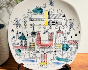 May include: A white ceramic plate with a hand-painted illustration of the Oslo skyline. The illustration features buildings, trees, a boat, and people. The text "OS LO" is written at the bottom of the plate.