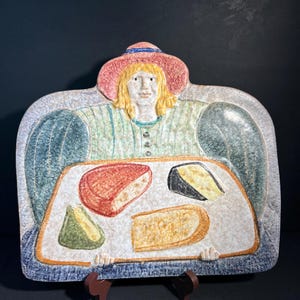 Puede incluir: Una fuente de cerámica que representa a una mujer con un sombrero rosa y una camisa verde a rayas. La fuente contiene cuatro cuñas de queso de colores. Los bordes son azules y el fondo es negro.