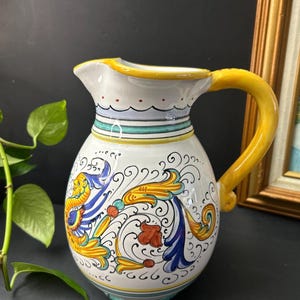 May include: A white ceramic jug with a yellow handle and rim. The jug is decorated with blue, yellow, and red floral patterns. The jug has a height of approximately 20 cm.