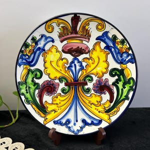 May include: A decorative ceramic plate with a vibrant floral design. The plate features a white background with a colourful pattern of yellow, blue, green, and purple floral motifs. The plate has a dark blue rim and is displayed on a wooden stand.