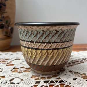 Może przedstawiać: Ceramiczna miska z teksturowanym, wzorzystym wzorem. Miska ma pasma kolorów, w tym brązowy, żółty i niebiesko-zielony. Obręcz jest ciemnobrązowa. Misa stoi na białej koronkowej serwetce.