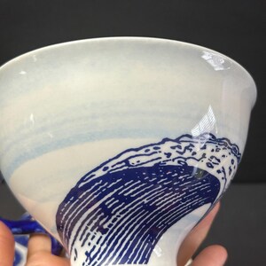May include: White ceramic bowl with a blue wave design. The bowl features a wide opening and a curved base. The wave design is a deep blue color and covers a portion of the bowl's exterior. The bowl has a glossy finish.