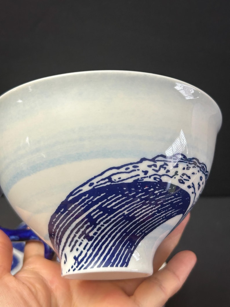 May include: White ceramic bowl with a blue wave design. The bowl features a wide opening and a curved base. The wave design is a deep blue color and covers a portion of the bowl's exterior. The bowl has a glossy finish.