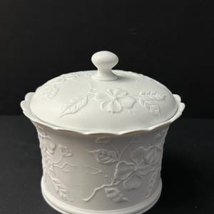 May include: A white ceramic jar with a lid. The jar and lid are decorated with embossed floral and leaf patterns. The lid has a small, round knob. The jar is cylindrical with a scalloped edge.