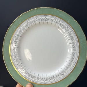 May include: A white plate with a pale green rim and gold trim. The plate features a decorative pattern and a scalloped edge. This plate is likely part of a dinner service.