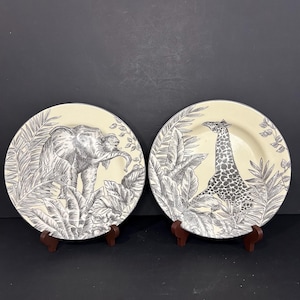 以下が含まれることがあります： アフリカの野生動物の白黒イラストが描かれた装飾用プレート2枚。1枚はゾウ、もう1枚はキリンが描かれており、どちらも熱帯の葉に囲まれています。プレートは小さな木製のスタンドに飾られています。