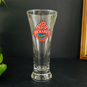 May include: A clear, tulip-shaped beer glass with the Rickard's Red logo in red, white, and blue. The glass has a flared top and a sturdy base. The logo features a stylized "R" and the words "Rickard's Red" in a circular design.