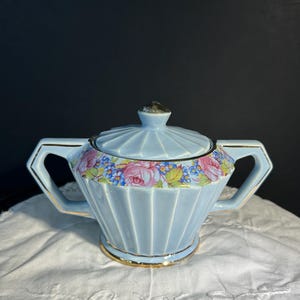 Peut inclure: Sucrier en céramique bleu clair avec couvercle, orné de roses roses et de fleurs bleues. Le sucrier a deux anses et une bordure dorée. Le couvercle est surmonté d'un petit bouton décoratif.