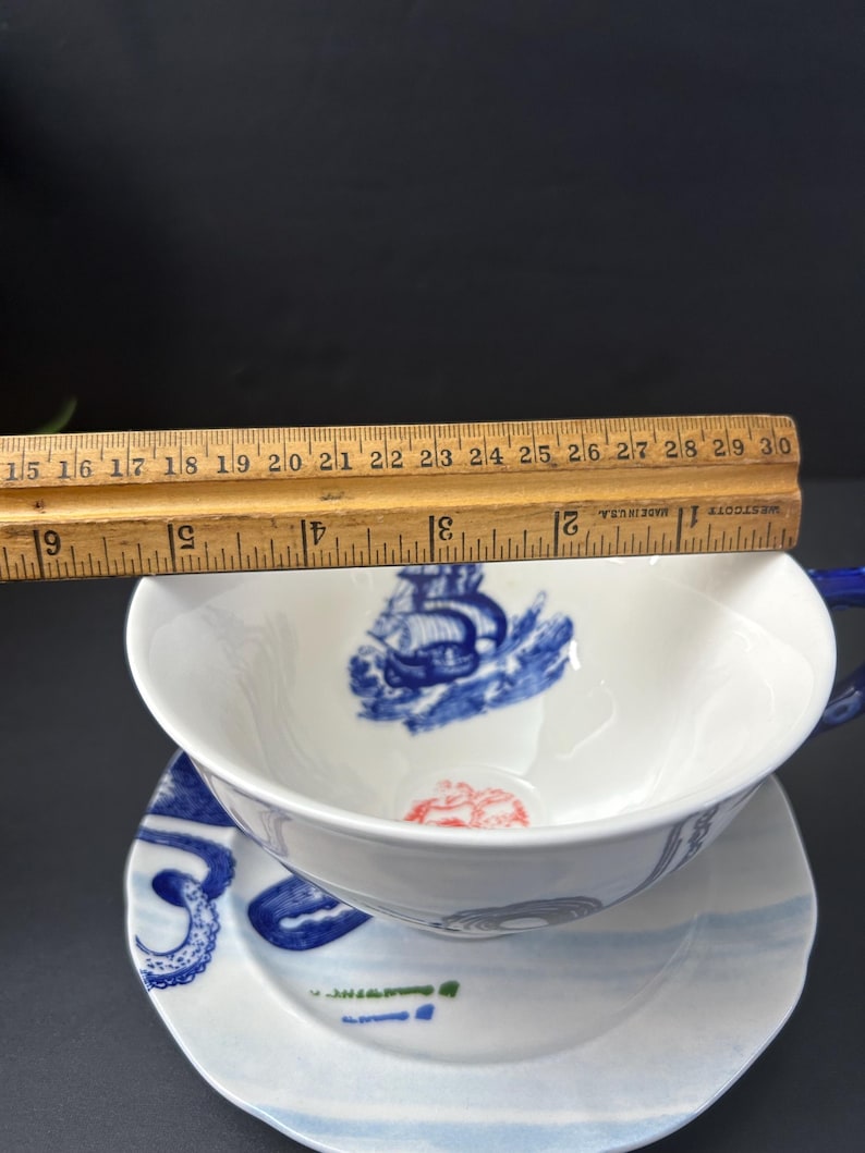 May include: White teacup and saucer set with blue nautical illustrations. The teacup is approximately 4 inches in diameter. The saucer has a matching design. The set includes a blue handle and rim. The design includes a ship and octopus.