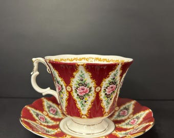 May include: A red teacup and saucer set with a floral design. The cup and saucer feature a repeating pattern of pink roses and green leaves on a white and gold background. The cup has a white handle and base.