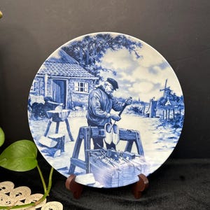 Könnte beinhalten: Blau-weiße Zierteller mit einer detaillierten Illustration eines Handwerkers, der an Holzschuhen arbeitet. Die Szene zeigt ein Haus, Werkzeuge und eine Windmühle im Hintergrund. Der Teller wird auf einem Holzständer präsentiert.