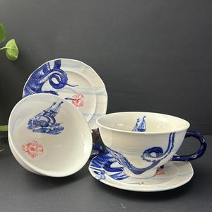 May include: A white ceramic teacup, saucer, and two plates with a blue nautical design. The design includes stylized waves, a sailboat, and a red floral accent. The teacup has a blue handle.