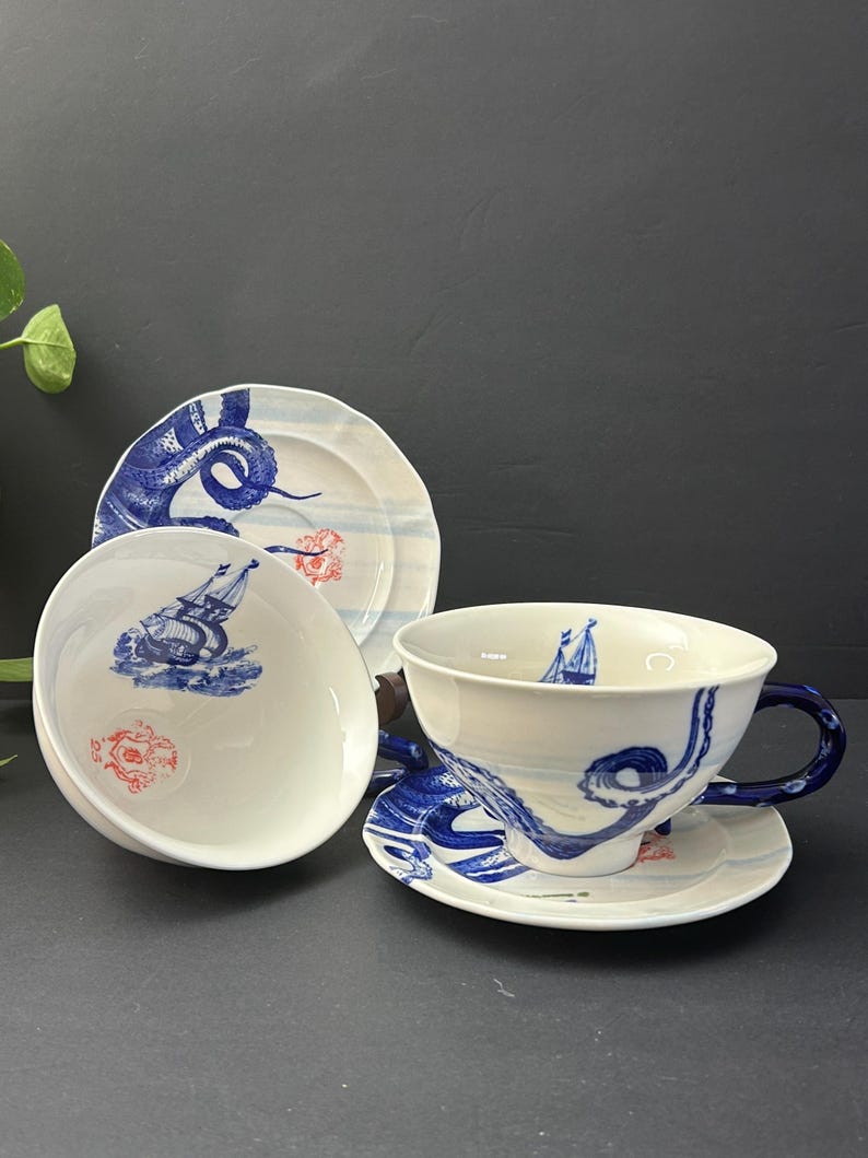 May include: A white ceramic teacup, saucer, and two plates with a blue nautical design. The design includes stylized waves, a sailboat, and a red floral accent. The teacup has a blue handle.
