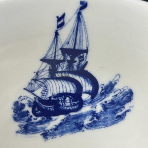 May include: White ceramic teacup with a blue illustration of a sailboat on the ocean. The ship has sails and flags, and the waves are depicted in detail. The design is a classic nautical theme.