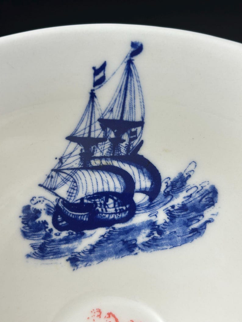 May include: White ceramic teacup with a blue illustration of a sailboat on the ocean. The ship has sails and flags, and the waves are depicted in detail. The design is a classic nautical theme.