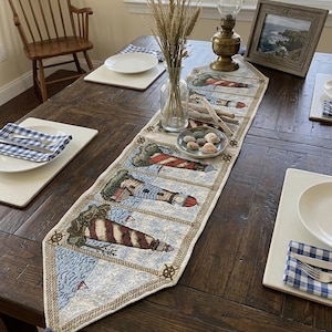 Op de afbeelding: Een decoratieve tafelloper met een nautisch vuurtorenontwerp in rood, wit en blauw. De loper ligt op een donkere houten tafel, aangevuld met witte borden, blauw-wit geruite servetten en een vintage olielamp.