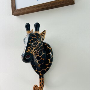 May include: A wooden giraffe head wall hook. The giraffe is painted black with brown spots and a brown and tan hook. The giraffe is mounted on a white wall, with a framed picture in the background.