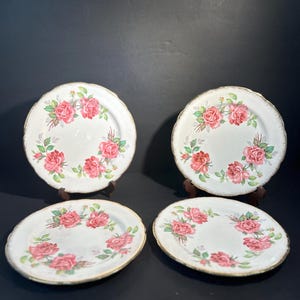 Può includere: Quattro piatti in ceramica bianca con un motivo floreale di rose rosa e bordi dorati. I piatti sono disposti su una superficie scura, mettendo in mostra il loro motivo decorativo. I piatti hanno bordi smerlati.
