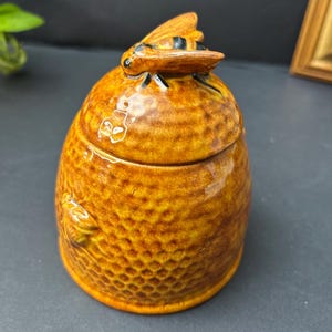 May include: A ceramic honey pot shaped like a beehive with a glossy amber glaze and a textured honeycomb pattern. The lid features a sculpted bee with black and yellow stripes resting on top, and a small bee is moulded into the side of the pot.
