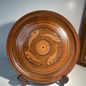 Puede incluir: Un plato de cerámica marrón con un diseño de peces. El plato presenta un patrón circular de peces alrededor de una espiral central. El plato se exhibe sobre un soporte de madera. El plato tiene aproximadamente 25 cm de diámetro.