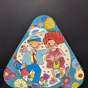 Könnte beinhalten: Eine dreieckige Keramikplatte mit einem skurrilen Design. Sie zeigt ein tanzendes Paar, umgeben von bunten Blumen. Die Platte hat einen türkisfarbenen Rand und einen weißen Innenraum mit Tupfen. Die Figuren sind in leuchtenden Farben gehalten, was die verspielte Ästhetik unterstreicht.