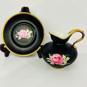 May include: A black and gold creamer and saucer set with a pink rose design. The creamer has a handle and a spout. The saucer is round and has a gold rim.
