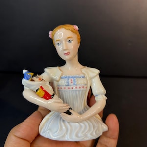 Puede incluir: Figura de porcelana de una mujer con un vestido blanco que sostiene una muñeca pequeña. La mujer tiene el pelo rubio con lazos rosas y lleva un corsé azul claro. La muñeca está vestida de rojo, azul y amarillo.