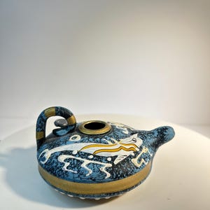 May include: A ceramic oil lamp with a blue, white, and gold design. The lamp features a handle and a spout. The body of the lamp is decorated with a white and yellow dragon-like figure.