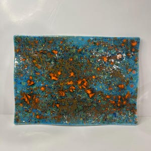 Puede incluir: Bandeja rectangular de cerámica con diseño moteado. La bandeja presenta un acabado brillante con un color base turquesa y motas naranjas y marrones vibrantes. Los bordes son ligeramente curvos, lo que añade un toque único al diseño.