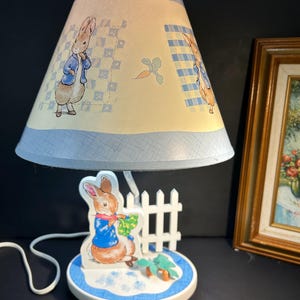 Puede incluir: Lámpara infantil con diseño de Peter Rabbit. La pantalla es azul claro y amarilla, decorada con ilustraciones de Peter Rabbit y zanahorias. La base presenta una figura 3D de Peter Rabbit, una valla blanca y modelos de zanahorias.