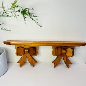 May include: A wooden shelf with two decorative bow-shaped brackets. The shelf is made of light brown wood and the brackets are a darker brown. The shelf is mounted on a white wall.