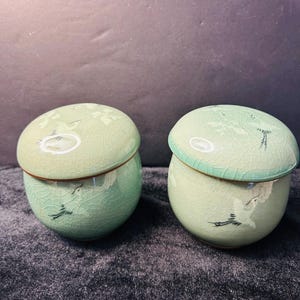 May include: Two celadon-colored ceramic jars with lids. Each jar has a round shape with a lid and a crackle glaze. The jars are decorated with a crane design in black. The jars are on a dark gray surface.