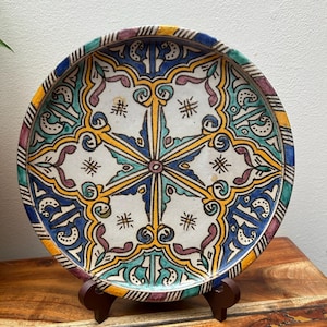 May include: A decorative ceramic plate with a detailed, symmetrical design. The plate has a white centre with a starburst pattern in yellow and blue, surrounded by teal, purple, and yellow accents. The rim has a striped pattern.