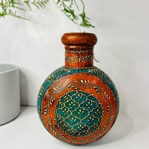 Può includere: Un vaso in ceramica dipinto a mano con intricati motivi floreali in tonalità arancio, turchese e oro. Il vaso ha un corpo rotondo e un collo stretto.
